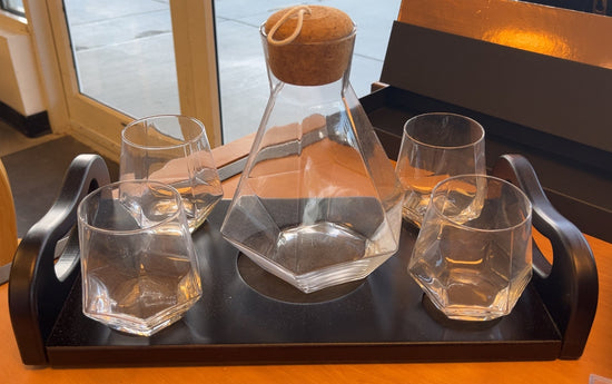 A clear glass carafe with a cork stopper and four matching faceted glass tumblers arranged on a mahogany serving tray, photographed on a warm wood table in a real indoor setting.